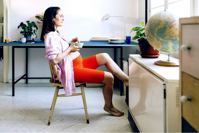 Eine schwangere Frau sitzt entspannt auf einem Holzstuhl in einem hellen Zimmer und hält eine Tasse und eine Schüssel in der Hand. Sie trägt leuchtend orangefarbene Shorts und ein hellrosa Oberteil. Ein Globus, Zimmerpflanzen und ein Schreibtisch mit verschiedenen Gegenständen schaffen eine gemütliche und ungezwungene Atmosphäre.