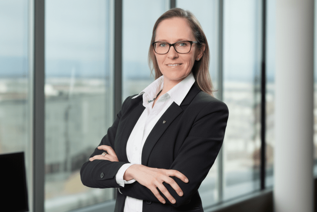 HBV Sabine Hufnagel steht mit verschränkten Armen in einem modernen Büro. Sie trägt einen schwarzen Hosenanzug und eine weiße Bluse und strahlt Selbstbewusstsein und Autorität aus. Große Fenster geben den Blick auf die urbane Skyline im Hintergrund frei, und die Atmosphäre ist formell und geschäftsmäßig. Das natürliche Licht trägt zu einem klaren und modernen Ambiente bei.