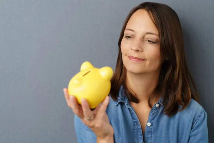 A person holds a bright yellow piggy bank in their hand, symbolizing savings or financial planning. The setting features a neutral gray background and casual attire, creating a relaxed and approachable mood. The piggy bank has a playful design, resembling a cartoonish animal.