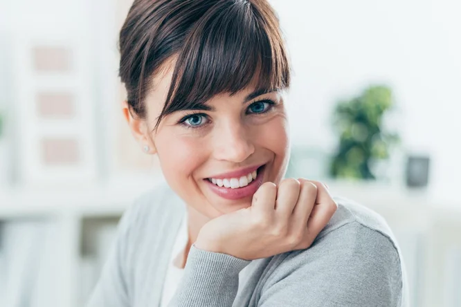 Eine Person sitzt in einem legeren Raum und trägt eine hellgraue Strickjacke. Im Hintergrund sind sanft verschwommene Elemente wie Grünpflanzen und Bücherregale zu sehen, die eine entspannte und zugleich professionelle Atmosphäre schaffen. Neutrale Farbtöne dominieren die Farbpalette und unterstreichen den ruhigen und zurückhaltenden Stil.