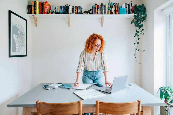 Eine junge Frau steht an einem Tisch in einem hellen, modernen Homeoffice und schreibt auf Papier, während sie gleichzeitig auf ihren Laptop schaut. Der Raum ist minimalistisch eingerichtet: weiße Wände, Holzmöbel und ein Bücherregal darüber. Die Atmosphäre ist konzentriert und produktiv; natürliches Licht strömt herein, und ein gestreiftes Hemd verleiht dem Outfit eine lässige Note. Weder Zahlen noch Marken sind sichtbar.