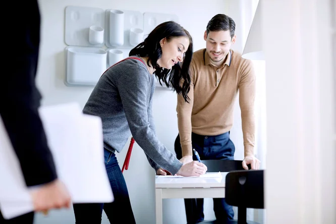 Zwei Personen führen in einem modernen Büro ein gemeinsames Gespräch. Eine Person schreibt auf einem Dokument, das auf einem weißen Tisch liegt, während eine andere danebensteht und zusieht. Der Hintergrund ist minimalistisch gestaltet und wird von sanftem Tageslicht durch ein nahes Fenster erhellt. Eine dritte Person ist teilweise sichtbar und hält Papiere in der Hand.