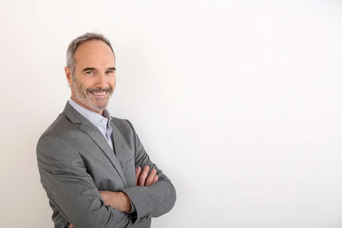 Ein Mann in einem grauen Anzug und hellem Hemd steht selbstbewusst mit verschränkten Armen vor einem schlichten weißen Hintergrund. Die minimalistische Gestaltung unterstreicht die professionelle Kleidung und Haltung. Das Bild vermittelt eine formelle und souveräne Stimmung und eignet sich daher für geschäftliche oder unternehmensbezogene Zwecke.