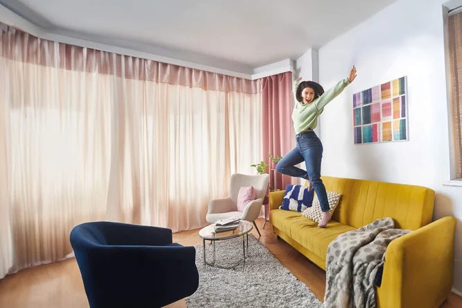 Eine junge Frau mit lockigem Haar springt vergnügt auf einem leuchtend gelben Sofa in einem modernen, hell erleuchteten Wohnzimmer. Der Raum ist mit pastellfarbenen Vorhängen, einem farbenfrohen Wandbild und gemütlichen Möbeln wie einem blauen Sessel und einem kleinen runden Couchtisch ausgestattet. Die Atmosphäre ist lebendig und verspielt, wobei die Mischung aus warmen und kühlen Farbtönen ein harmonisches Gesamtbild ergibt.