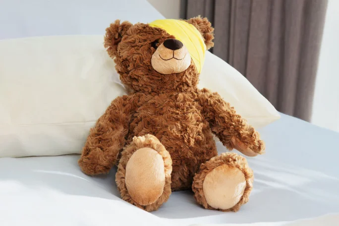 Ein weicher, brauner Teddybär sitzt auf einem weißen Bett, im Hintergrund ist ein Kissen zu sehen. Um seinen Kopf ist ein gelber Verband gewickelt, der auf Pflege oder Genesung hindeutet. Die Szenerie ist hell und minimalistisch, die neutralen Farbtöne schaffen eine behagliche Atmosphäre.