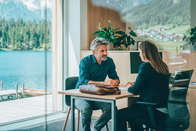Zwei Erwachsene unterhalten sich an einem modernen Schreibtisch. Durch große Fenster bietet sich ein malerischer Blick auf See und Berge. Der Arbeitsplatz ist lichtdurchflutet, mit Zimmerpflanzen ausgestattet und minimalistisch eingerichtet. Beide sind professionell gekleidet, was auf ein formelles Geschäftsumfeld hindeutet. Die Atmosphäre ist ruhig und konzentriert, untermalt von den natürlichen Grün- und Blautönen der umliegenden Landschaft.