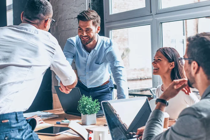 Eine professionelle Arbeitsumgebung mit einer Gruppe von Kollegen in einem modernen Büro. Zwei Personen geben sich die Hand, ein Symbol für Zusammenarbeit oder Einigung. Der Arbeitsplatz ist mit Laptops, Dokumenten und einer kleinen Topfpflanze ausgestattet; durch große Fenster strömt natürliches Licht.