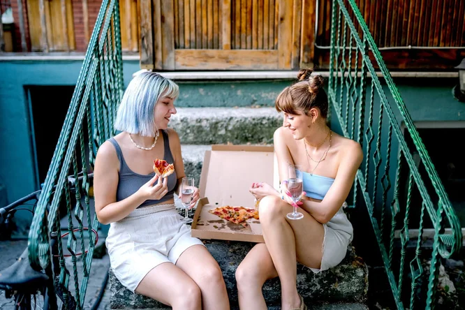 Zwei junge Frauen sitzen auf einer Außentreppe und teilen sich eine Pizza aus einem offenen Karton. Sie halten Gläser mit Wasser oder anderen klaren Getränken in der Hand, was eine ungezwungene und entspannte Atmosphäre schafft. Im Hintergrund sind grüne Geländer und Holztüren zu sehen, die auf ein urbanes oder wohnliches Umfeld hindeuten.