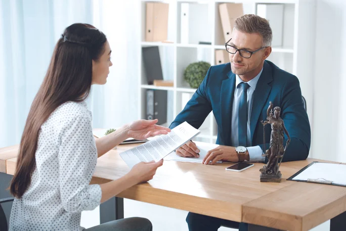 Ein formelles Treffen zweier Personen in einem hellen, modernen Büro. Auf dem Tisch liegen Dokumente, ein Smartphone und eine dekorative Justitia-Statue. Regale im Hintergrund beherbergen Bücher und Büromaterialien und unterstreichen die professionelle Atmosphäre.