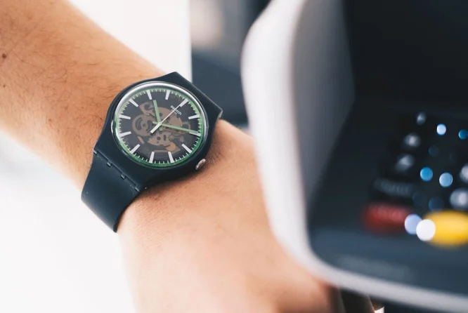 Eine Nahaufnahme zeigt das Handgelenk einer Person mit einer schwarzen analogen Armbanduhr mit grünen Akzenten und sichtbarem Uhrwerk. Die Person hält ihre Hand in der Nähe eines Zahlungsterminals, was auf eine Transaktion oder kontaktloses Bezahlen hindeutet. Der Hintergrund ist leicht verschwommen, wodurch der Fokus auf der Uhr und der Handlung liegt. Das Zifferblatt der Uhr hat ein modernes Skelett-Design. Weder auf der Uhr noch auf dem Terminal sind Schriftzüge oder Zahlen sichtbar.