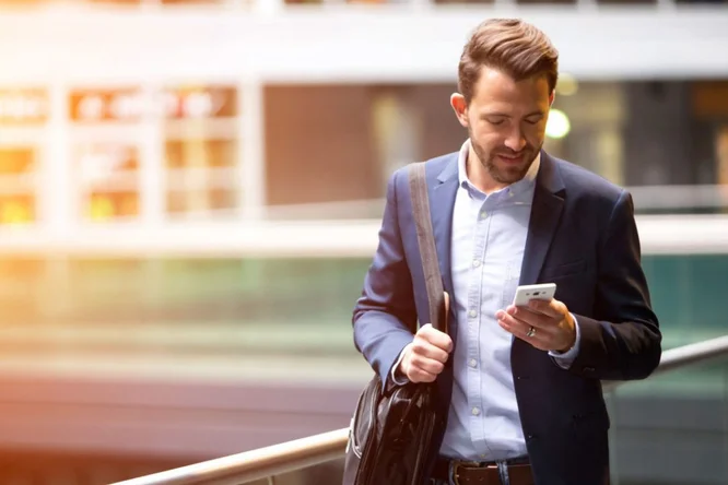 Ein junger Mann in formeller Kleidung hält in einer modernen Stadtumgebung ein Smartphone in der Hand. Er trägt eine schwarze Umhängetasche und scheint in sein Gerät vertieft zu sein. Im Hintergrund bilden Glasarchitektur und warmes Sonnenlicht eine Atmosphäre, die professionell und gleichzeitig ungezwungen wirkt.