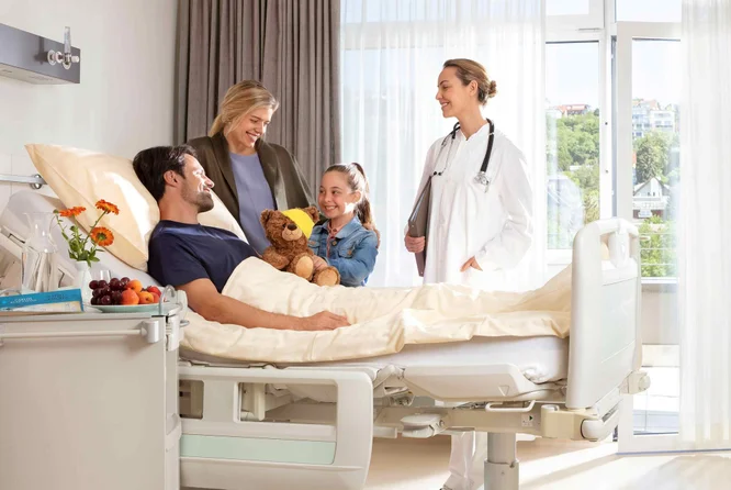 unge Familie besucht den Vater im Krankenhauszimmer, während eine Ärztin lächelnd zusieht.
