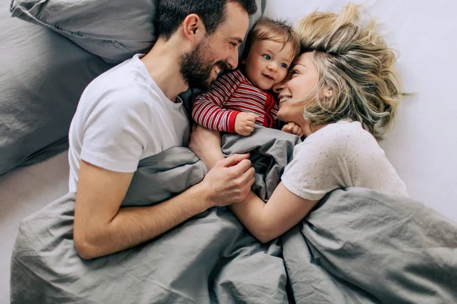 Eine Familie liegt gemütlich im Bett, eingehüllt in weiche, graue Bettwäsche. Die Atmosphäre ist behaglich und intim, entspannt und entspannt. Das Kind trägt ein rot-weiß gestreiftes Outfit, das einen Farbtupfer in die neutrale Farbpalette setzt.