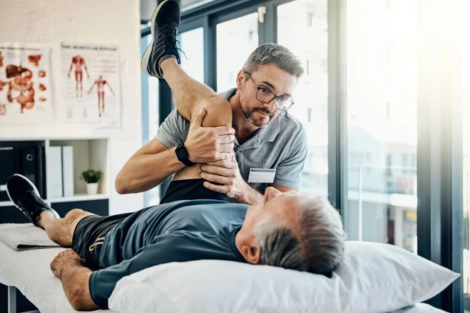 Ein älterer Mann liegt während einer Physiotherapie-Sitzung in einer hellen, modernen Klinik auf einer Behandlungsliege. Der Therapeut hilft ihm bei einer Beindehnung und konzentriert sich dabei auf Beweglichkeit und Rehabilitation. Anatomische Tafeln und große Fenster im Hintergrund unterstreichen die professionelle Atmosphäre.
