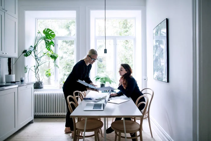 Zwei Frauen unterhalten sich an einem weißen Esstisch in einer hellen, modernen Küche. Der Raum ist lichtdurchflutet dank großer Fenster, Zimmerpflanzen und minimalistischer Einrichtung. Ein Laptop und Dokumente liegen auf dem Tisch und deuten auf eine Arbeits- oder Lernsitzung hin.