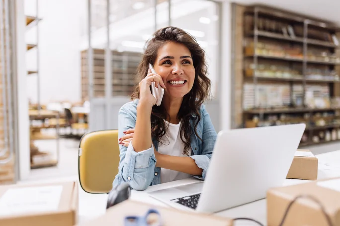 Cheerful businesswoman speaking on the phone while working in a warehouse. Happy online store owner making plans for product shipping. Female entrepreneur running an e-commerce small business.