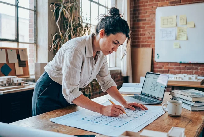 Eine Frau konzentriert sich an einem Holzschreibtisch in einem hellen, industriell gestalteten Arbeitszimmer auf das Zeichnen von Architekturplänen. Große Fenster, freigelegte Backsteinwände und ein Laptop mit Grundrissen prägen den Raum. Die Atmosphäre ist kreativ und professionell, natürliches Licht und Erdtöne dominieren. Weder Zahlen noch Marken sind sichtbar.
