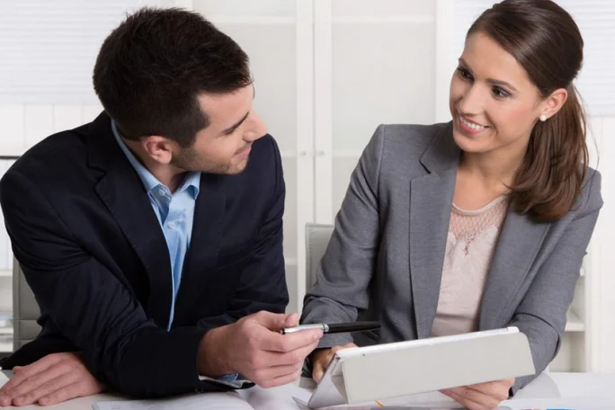 Two business professionals are seated at a desk in a modern office setting, engaged in a discussion while using a tablet device. The man is holding a pen, pointing towards the tablet, while the woman attentively listens. Both are dressed in formal business attire, suggesting a professional environment.