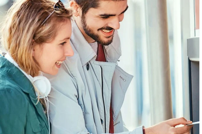 A man and a woman are interacting with an ATM machine in an outdoor setting. The woman is wearing headphones and a green jacket, while the man is dressed in a light-colored coat. The scene suggests a casual urban environment with natural lighting.