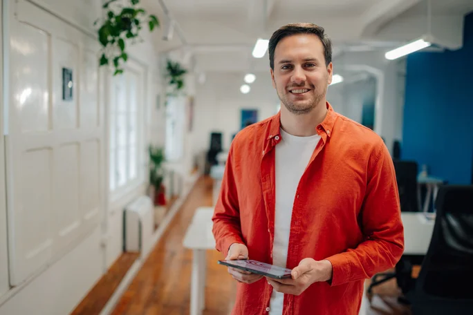 Startup founder wearing an orange shirt, holding a tablet and smiling confidently in a modern office corridor adorned with plants and featuring bright white and blue walls