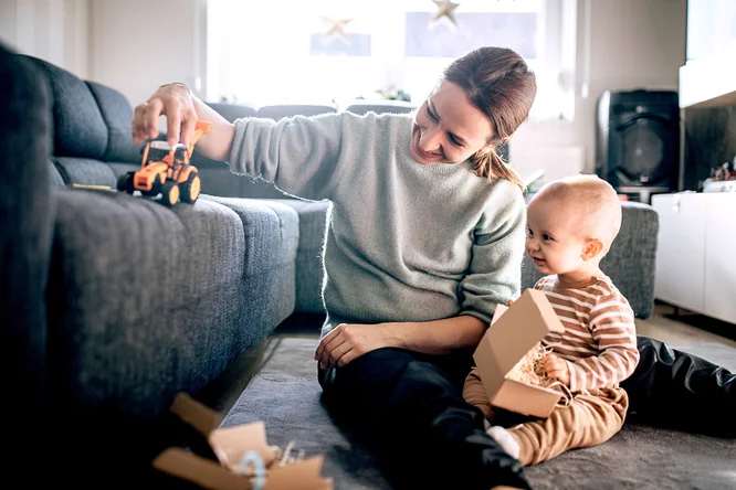 Ein Elternteil und ein kleines Kind sitzen in einem gemütlichen Wohnzimmer auf dem Boden und spielen mit einem Spielzeuglaster und Pappkartons. Der Raum ist hell und freundlich, da viel Tageslicht durch die Fenster strömt und eine warme, einladende Atmosphäre schafft. Das Kind trägt einen gestreiften Schlafanzug, das Elternteil ist leger in Pullover und Hose gekleidet.