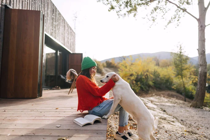 Eine junge Frau mit grüner Mütze und rotem Pullover sitzt mit einem aufgeschlagenen Buch auf einer Holzterrasse. Sie unterhält sich liebevoll mit einem weißen Hund in einer idyllischen, von Bäumen und Hügeln umgebenen Landschaft. Das Bild vermittelt durch das natürliche Licht und die herbstlichen Farbtöne eine friedliche und entspannte Stimmung.