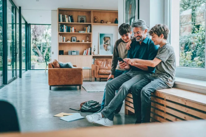 Ein Erwachsener und zwei Kinder sitzen auf einer Holzbank in einem geräumigen, modernen Wohnzimmer. Sie sind mit Smartphones beschäftigt, was auf einen Moment der Verbundenheit oder des Lernens hindeutet. Große Fenster, Bücherregale und ein gemütliches braunes Sofa prägen den Raum. Auf dem Boden liegen Papiere und ein Rucksack. Die Atmosphäre ist warm und entspannt, dank natürlichem Licht und neutralen Farbtönen.