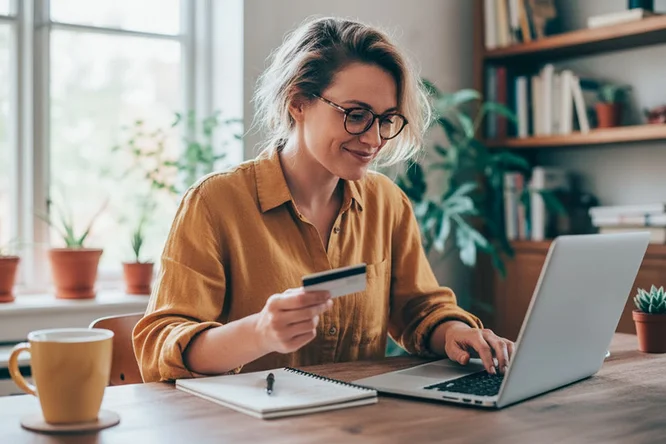 Eine Frau sitzt an einem Schreibtisch und arbeitet mit einem Laptop. In der Hand hält sie eine Kreditkarte, was auf Online-Shopping oder -Zahlungen hindeutet. Die Szenerie ist ein gemütliches Homeoffice mit Topfpflanzen und Tageslicht. Sie trägt ein senfgelbes Hemd und hat ein Notizbuch neben sich liegen – eine produktive und entspannte Atmosphäre. Weder Zahlen noch Marken sind sichtbar.