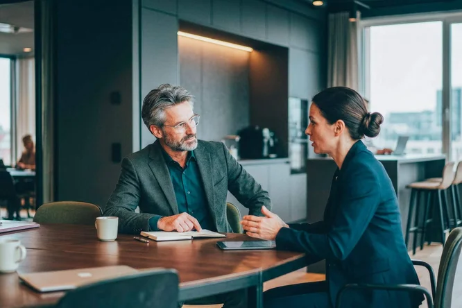 Zwei Erwachsene unterhalten sich an einem runden Tisch in einem modernen Büro. Der Arbeitsbereich zeichnet sich durch große Fenster, warmes Licht und minimalistische Einrichtung aus. Beide tragen Businesskleidung, was auf ein formelles oder kooperatives Meeting hindeutet. Kaffeetassen und Notizbücher auf dem Tisch unterstreichen die professionelle Atmosphäre. Die Stimmung ist konzentriert und professionell.