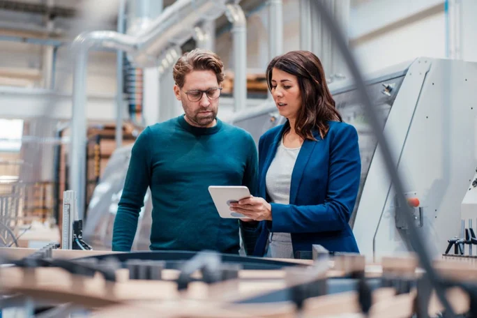 Zwei Personen stehen in einer modernen Fabrikhalle und prüfen Informationen auf einem Tablet. Die Umgebung ist mit Industriemaschinen und Rohrleitungen ausgestattet, was auf eine Produktionsstätte hindeutet. Die Personen scheinen in ein fachliches Gespräch oder eine Inspektion vertieft zu sein.