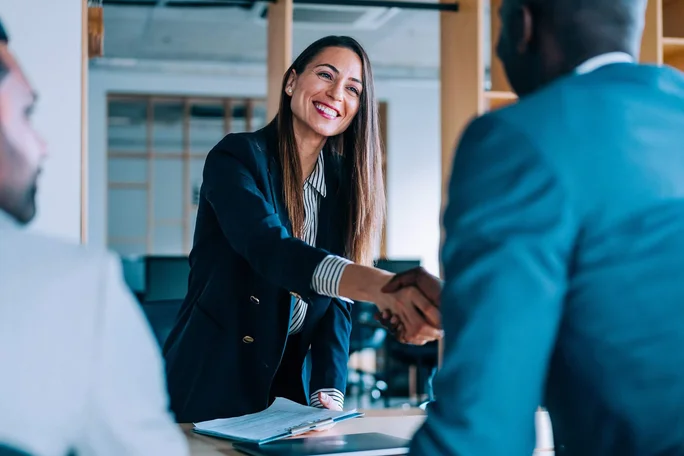 Eine berufstätige Frau in formeller Kleidung schüttelt einem Mann in einem modernen Büro die Hand. Der Raum ist mit Schreibtischen, Glastrennwänden und einem Gemeinschaftsarbeitsbereich ausgestattet. Das Bild vermittelt Professionalität, Teamwork und unternehmerisches Engagement.