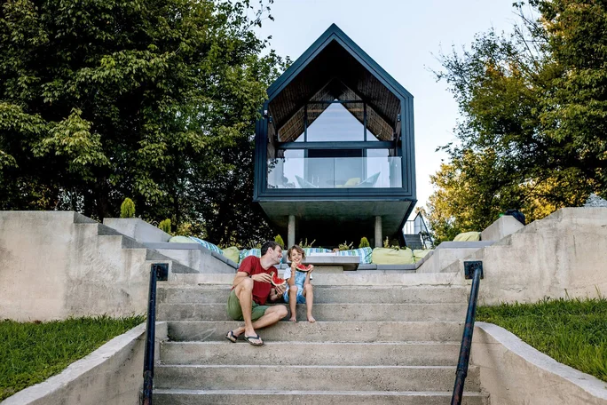 Ein modernes Haus mit dreieckigem Dach, umgeben von üppigem Grün. Eine dreiköpfige Familie – zwei Erwachsene und ein Kind – sitzt auf den Betonstufen, die zum Haus führen. Die Atmosphäre im Freien ist entspannt und ungezwungen, geprägt von klaren architektonischen Linien und natürlichen Elementen.