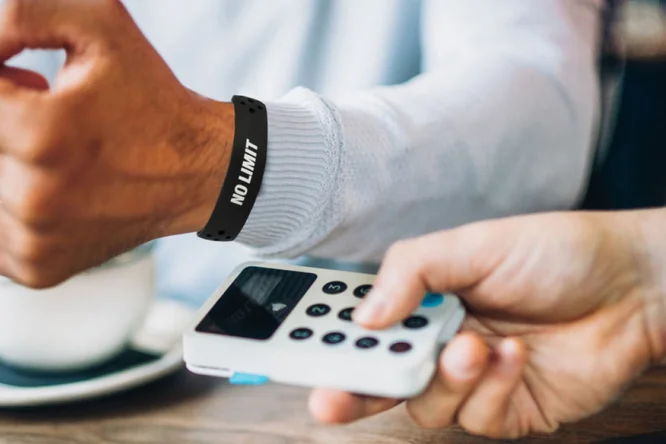 Eine Person mit einem schwarzen Armband mit der Aufschrift „NO LIMIT“ bezahlt kontaktlos mit einem mobilen Kartenlesegerät. Die Szenerie wirkt ungezwungen, möglicherweise in einem Café; im Hintergrund ist eine Tasse zu sehen. Der Fokus liegt auf der Transaktion und dem Aufdruck auf dem Armband.