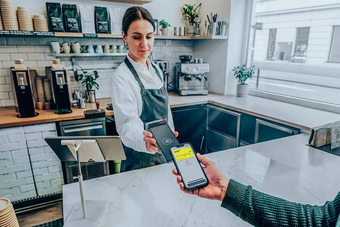 Ein Barista in Schürze steht hinter der Theke eines modernen Cafés, während ein Kunde sein Smartphone für eine kontaktlose Zahlung hochhält. Auf dem Display erscheint eine Zahlungsbestätigung mit gelber Akzentuierung und dem deutlich sichtbaren Betrag von 12,00 EUR. Das Café ist minimalistisch gestaltet, im Hintergrund sind Kaffeemaschinen und Topfpflanzen zu sehen.