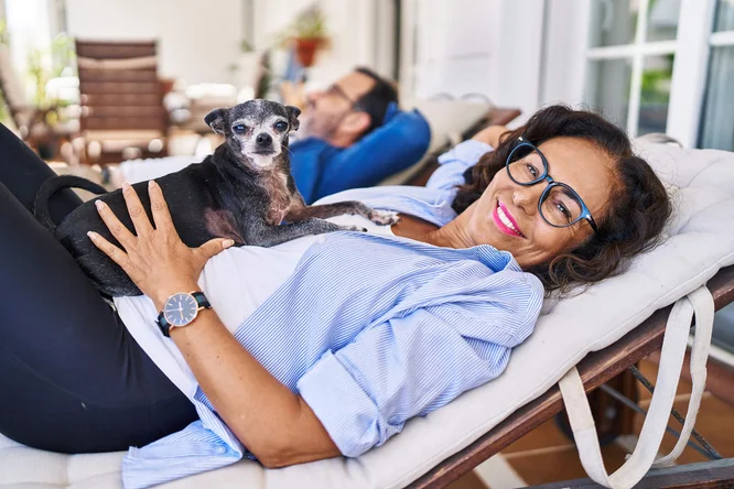 Ein kleiner, schwarz-grauer Hund ruht sich auf einer Person aus, die im Freien auf einer Liege liegt. Die Szene vermittelt eine entspannte Atmosphäre; im Hintergrund sind Gartenmöbel und Grünpflanzen zu sehen. Die Person trägt ein hellblaues Hemd und eine Armbanduhr, was dem lässigen Stil dezente Details verleiht.