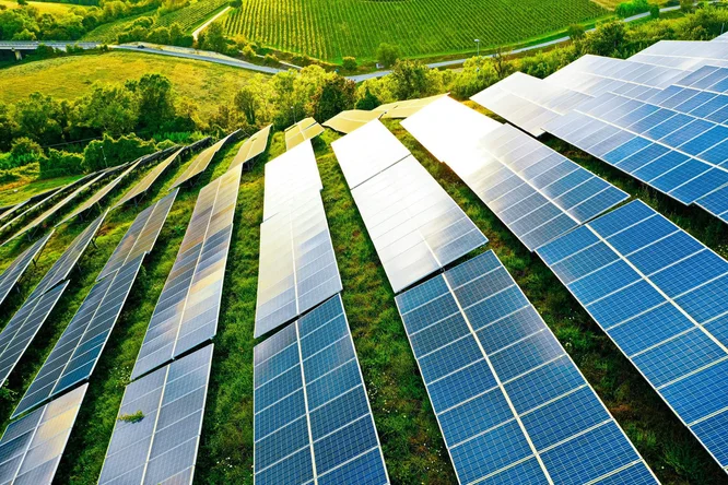 Eine große Solaranlage wurde auf einer Wiese inmitten von üppigem Grün und Ackerland installiert. Die Anlage demonstriert die Nutzung erneuerbarer Energien im ländlichen Raum. Helles Sonnenlicht lässt die reflektierenden Oberflächen der Paneele glänzen und unterstreicht so die Nachhaltigkeit und umweltfreundlichen Energielösungen.