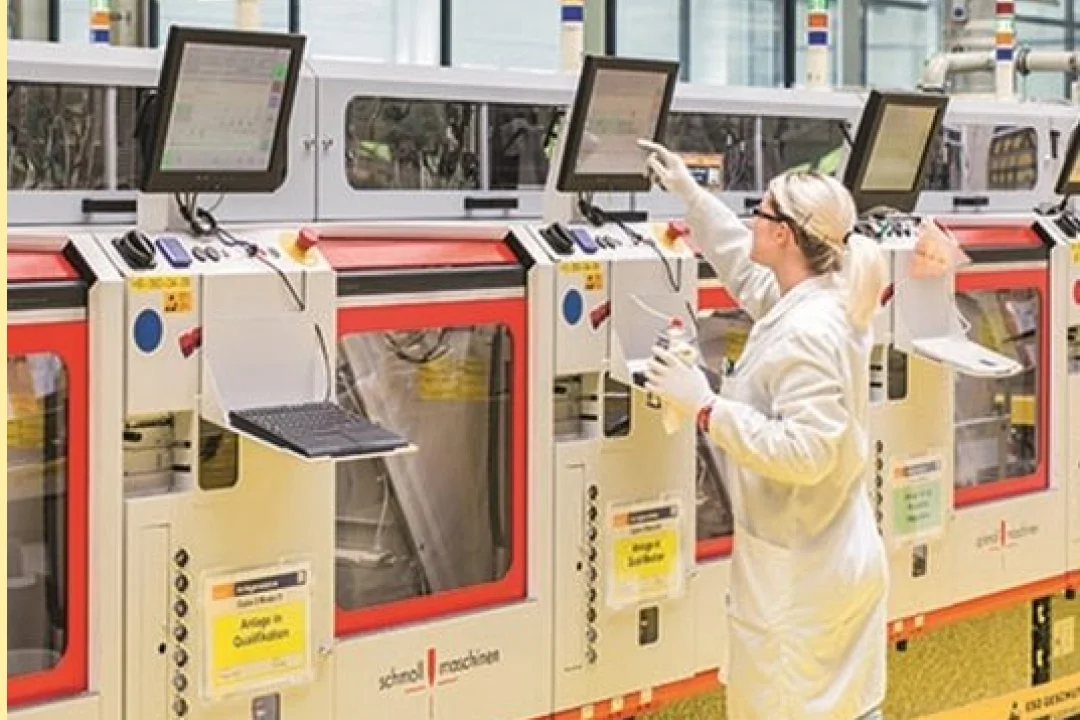 Ein Techniker im weißen Laborkittel bedient in einer Fabrikhalle moderne Industriemaschinen. Die Maschinen verfügen über mehrere Bildschirme, Bedienfelder und gut sichtbare Beschriftungen, darunter „Schnellmaschine“. Die Umgebung ist sauber und ordentlich und unterstreicht Präzision und Technologie in den Fertigungsprozessen.