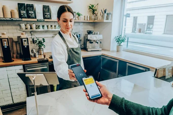 Ein Barista in Schürze hält ein kabelloses Kartenlesegerät an der Theke, während ein Kunde kontaktlos mit seinem Smartphone bezahlt. Das Café ist modern und hell eingerichtet; im Hintergrund sind Kaffeemaschinen, Tassen und Pflanzen zu sehen. Auf dem Smartphone-Bildschirm wird die Zahlungsbestätigung mit gelben Akzenten angezeigt. Die Atmosphäre ist entspannt und effizient, der Raum wird von natürlichem Tageslicht erhellt.