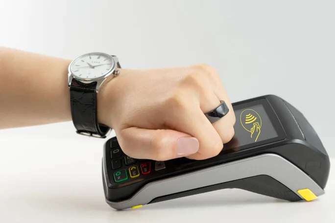 Nahaufnahme einer Hand mit einem schwarzen Smartring, die kontaktlos an einem Kartenlesegerät bezahlt. Die minimalistische Darstellung vor weißem Hintergrund betont die Technologie. Das Kartenlesegerät zeigt ein gelbes Symbol für kontaktloses Bezahlen an, das die Transaktionsbereitschaft signalisiert.