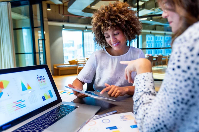 Zwei Personen arbeiten in einem modernen Büro zusammen und besprechen Datenvisualisierungen auf Laptop und Tablet. Der Arbeitsplatz mit großen Fenstern und zeitgemäßer Einrichtung vermittelt ein professionelles Ambiente. Diagramme, Grafiken und numerische Daten werden auf den Bildschirmen angezeigt und veranschaulichen Analysen und Leistungskennzahlen.