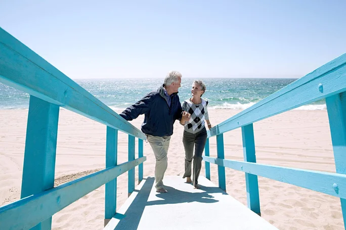 Ein älteres Paar spaziert Hand in Hand auf einer hellblauen Holzrampe, die zu einem Sandstrand führt. Die Meereswellen und der klare Himmel schaffen eine friedliche und sonnige Atmosphäre. Die Szenerie lädt zu entspannten Aktivitäten am Meer ein.
