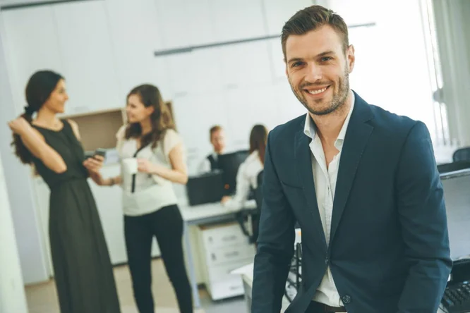 Ein Mann im dunkelblauen Anzug sitzt im Vordergrund eines modernen Büros. Im Hintergrund unterhalten sich drei Kollegen an Schreibtischen und Computern. Die Umgebung zeichnet sich durch helle Beleuchtung und eine professionelle Atmosphäre aus, in der Zusammenarbeit und Teamwork im Vordergrund stehen.