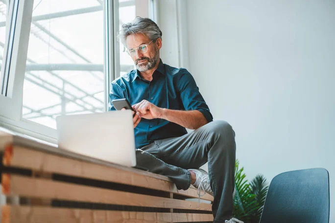 Ein Mann sitzt lässig auf einer Holzbank am Fenster und nutzt sein Smartphone, während vor ihm ein Laptop steht. Es wirkt wie ein moderner Arbeitsplatz mit viel Tageslicht. Grüne Pflanzen und minimalistische Möbel unterstreichen die zeitgemäße Atmosphäre.