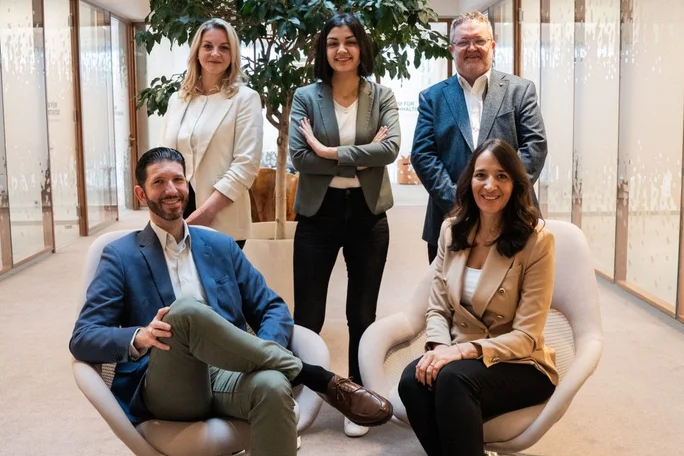Eine Gruppe von fünf Personen in Businesskleidung sitzt und steht in einem modernen Büro. Die Räumlichkeiten zeichnen sich durch Glaswände, eine große Zimmerpflanze und Möbel in neutralen Farbtönen aus. Die Atmosphäre vermittelt Professionalität und Zusammenarbeit und unterstreicht den Fokus auf Teamwork und ein professionelles Arbeitsumfeld.