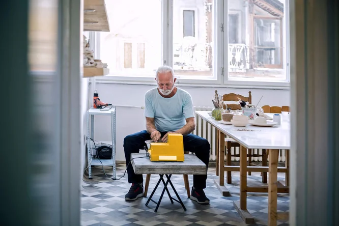 Ein Mann sitzt an einem kleinen Tisch und tippt auf einer leuchtend gelben Schreibmaschine in einem hellen Raum mit großen Fenstern. Die Szenerie wirkt wie ein gemütliches, modernes Zuhause mit Holzmöbeln und gemustertem Boden. Im Hintergrund sieht man einen Esstisch mit Pflanzen und Geschirr, was eine ungezwungene und kreative Atmosphäre schafft.