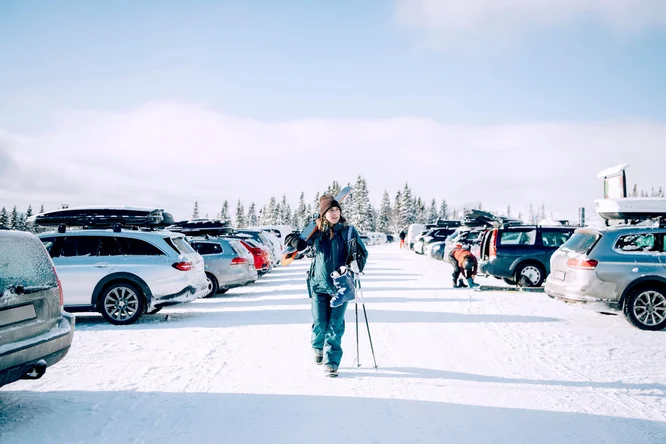Eine Frau in Winterkleidung trägt Skier und Stöcke über einen schneebedeckten Parkplatz. Im Hintergrund sind geparkte Autos mit Skiträgern und schneebedeckte Bäume unter strahlend blauem Himmel zu sehen. Die Szene vermittelt die Atmosphäre einer Wintersportaktivität in einer idyllischen Naturlandschaft.