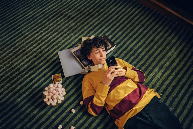 Eine Person in einem gelb-rot gestreiften Pullover liegt auf einem grün gestreiften Teppich und hält ein Smartphone in der Hand. Daneben liegen Bücher, ein kleiner Haufen Marshmallows und ein Polaroidfoto. Die Szenerie vermittelt eine gemütliche und entspannte Atmosphäre, in der Freizeit und Komfort im Vordergrund stehen.