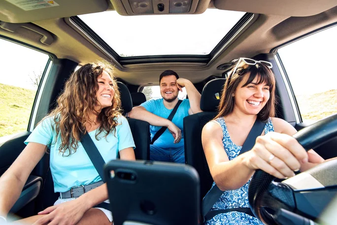 Drei Personen sitzen in einem Auto mit Schiebedach und genießen einen Roadtrip. Die Szene zeigt einen hellen, sonnigen Tag mit klarem Himmel, der durch das Schiebedach sichtbar ist. Ein Smartphone ist in der Nähe des Armaturenbretts befestigt und dient vermutlich der Navigation oder Unterhaltung während der Fahrt. Der Innenraum ist modern gestaltet und die Sicherheitsgurte sind angelegt, was die Sicherheit unterstreicht.
