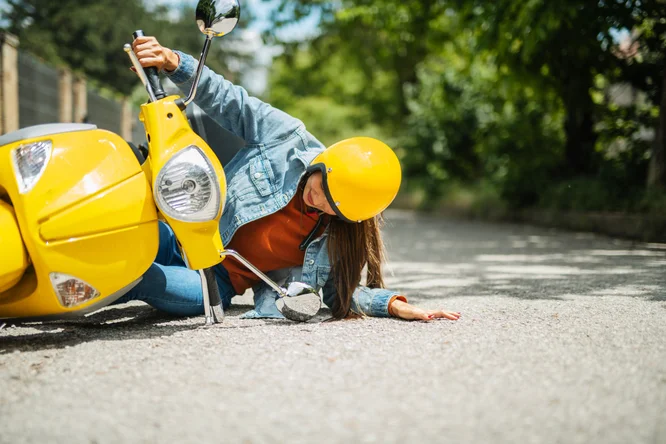 Eine junge Frau mit gelbem Helm und Jeansjacke stürzte auf einer asphaltierten Straße von einem leuchtend gelben Roller. Die Szene spielt im Freien, im Hintergrund ist Grün zu sehen. Der Seitenspiegel des Rollers ist zerbrochen, und die Frau scheint sich von dem Sturz zu erholen.