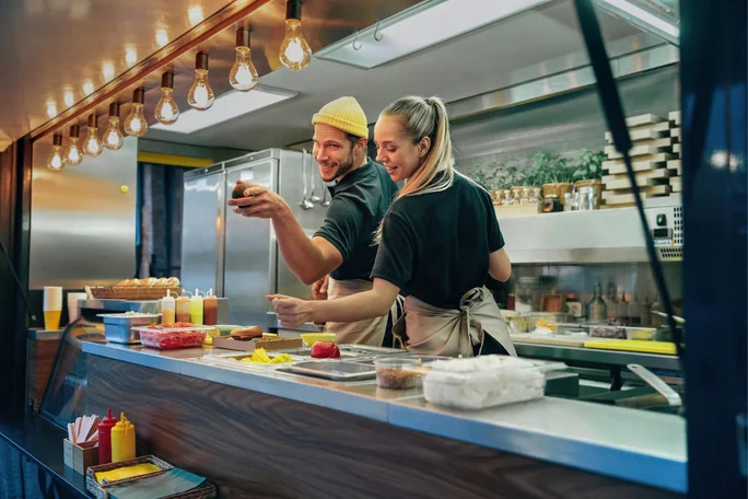 Zwei Köche bereiten in einem modernen Foodtruck Gerichte aus frischen Zutaten zu. Die Küche ist hell erleuchtet und mit sichtbaren Gewürzen, Tabletts und einer Holztheke ausgestattet. Warme Dekoleuchten sorgen für eine gemütliche Atmosphäre. Der Foodtruck verfügt über Edelstahlgeräte und übersichtliche Regale.